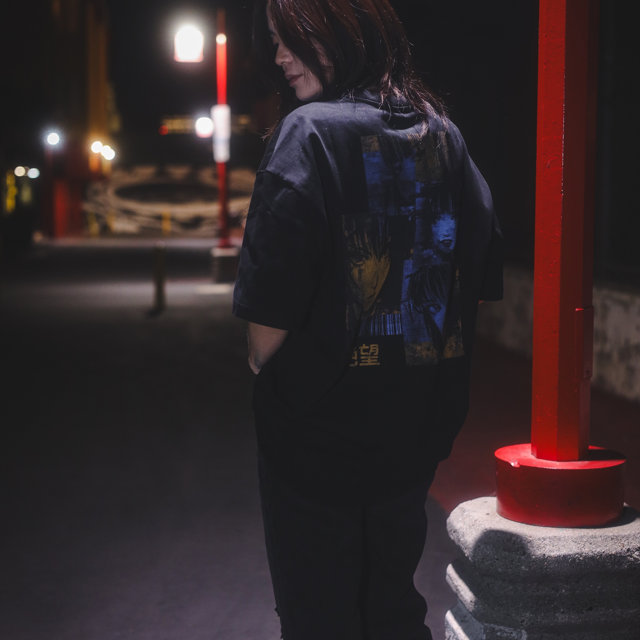 A person wearing a black oversized t-shirt with a blue and orange graphic on the back, standing next to a light pole at night.