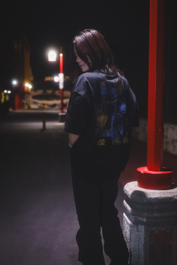 A person wearing a black oversized t-shirt with a blue and orange graphic on the back, standing next to a light pole at night.