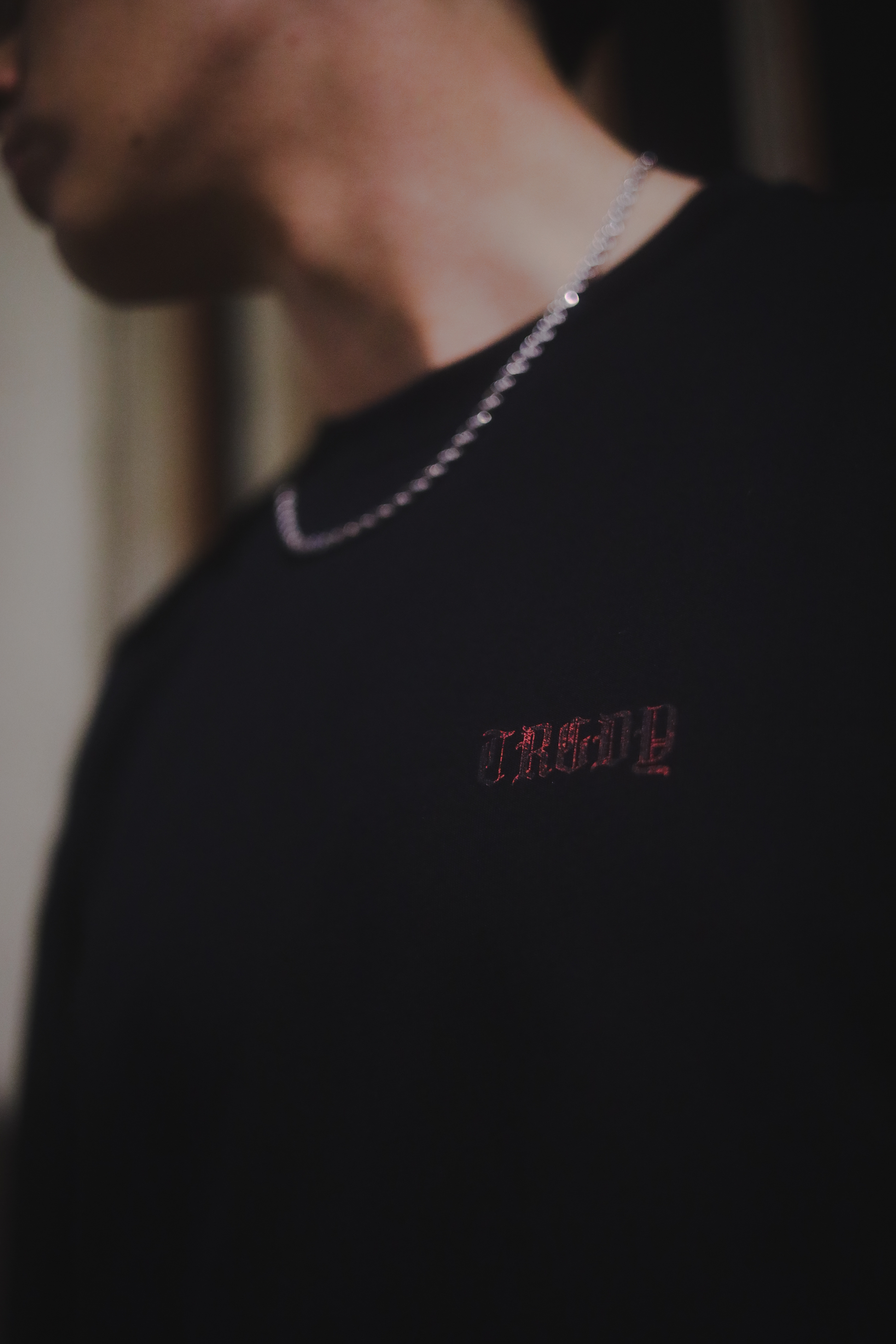 A person wearing a black oversize tee with a left side chest print in red gothic text that says "TRGDY". Person is standing against a dark background.