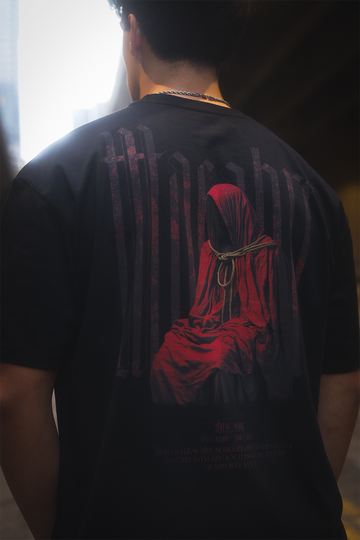 A close up of a person wearing a black oversize tee with a graphic of a hooded sitting figure with no face draped in a red cloak with a rope wrapped around the neck. With large stretched red gothic text that says "Macabre" with text underneath talking about what macabre is. Person is standing under a bridge in a city.