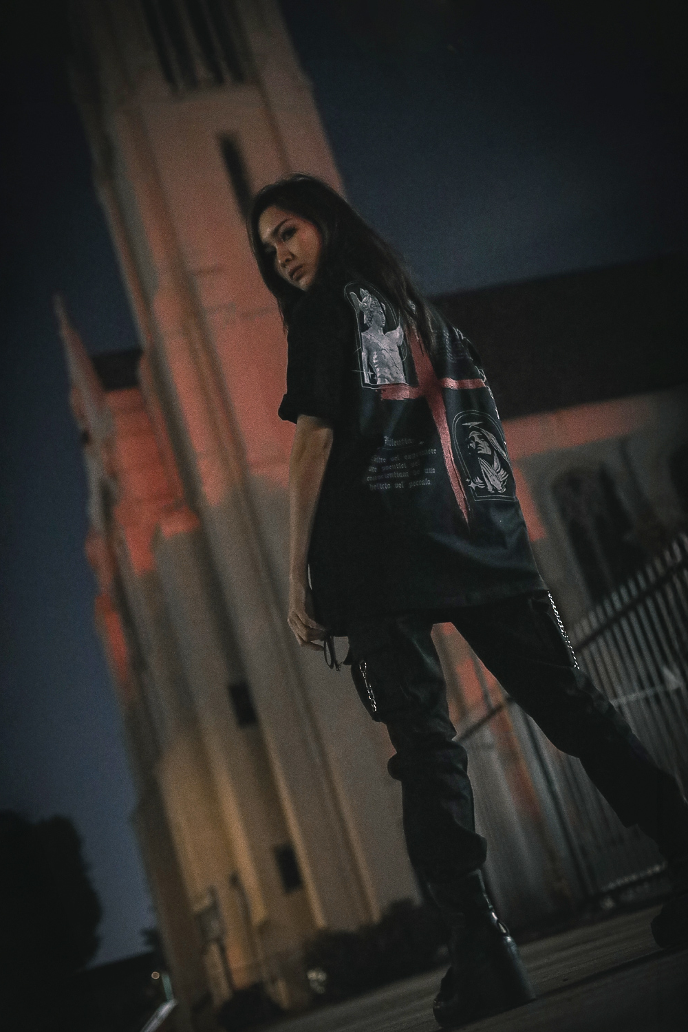 A person wearing a black oversize tee with a red cross on the back of her shirt, standing in front of a church at night