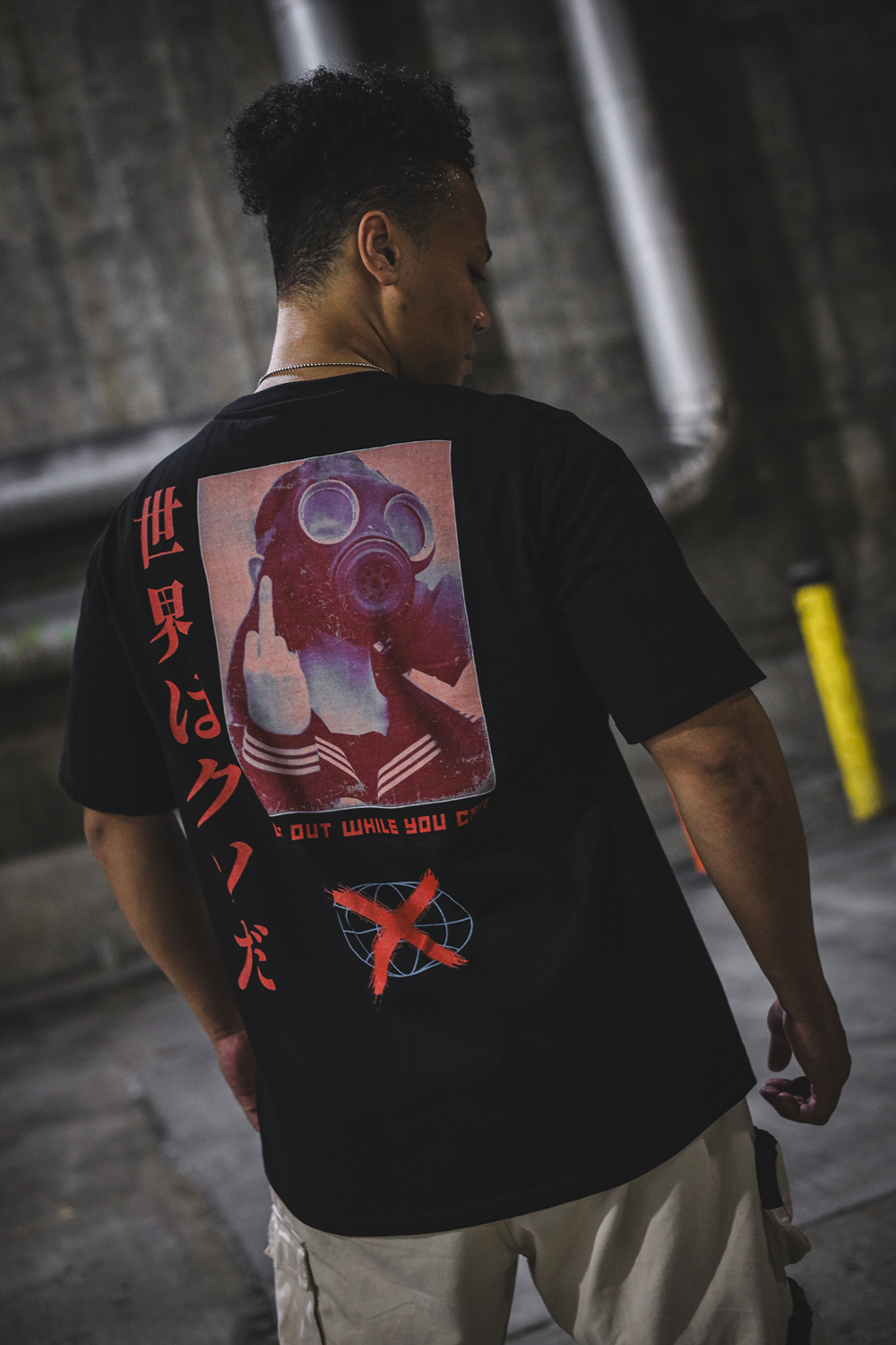 A person standing in a dark industrial setting wearing a black oversize tee with rear graphic of a reddish colored image of a girl in a gas mask doing a middle finger with orange/red text below stating "get out while you can". Left of the girl is a vertical orange/red text in Japanese saying "the world is sh*t". Bottom of the shirt is a bluish globe icon with a large orange/red X.