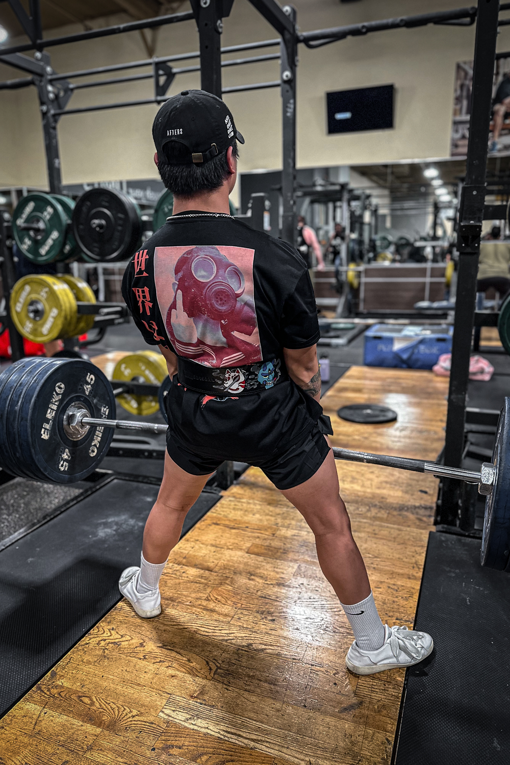 A person deadlifting heavy weight in the gym wearing a black oversize tee with a rear graphic of a reddish colored image of a girl in a gas mask doing a middle finger with orange/red text below stating "get out while you can". Left of the girl is a vertical orange/red text in Japanese saying "the world is sh*t". Bottom of the shirt is a bluish globe icon with a large orange/red X.