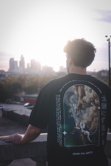 A person standing on a balcony overlooking a cityscape wearing a black oversize tee with a graphic of a hand reaching down from heaven to grab a falling man with wings. The left of the graphic talks about Icarus flying too close to the sun, there is also curved top text that says "don't get carried away with the infatuation of innovation and technology" and text to the right in Japanese that says "too close to the sun". Bottom print says "TRGDY EST: MMXXIV" The prints are in white. The tragedy of Icarus.