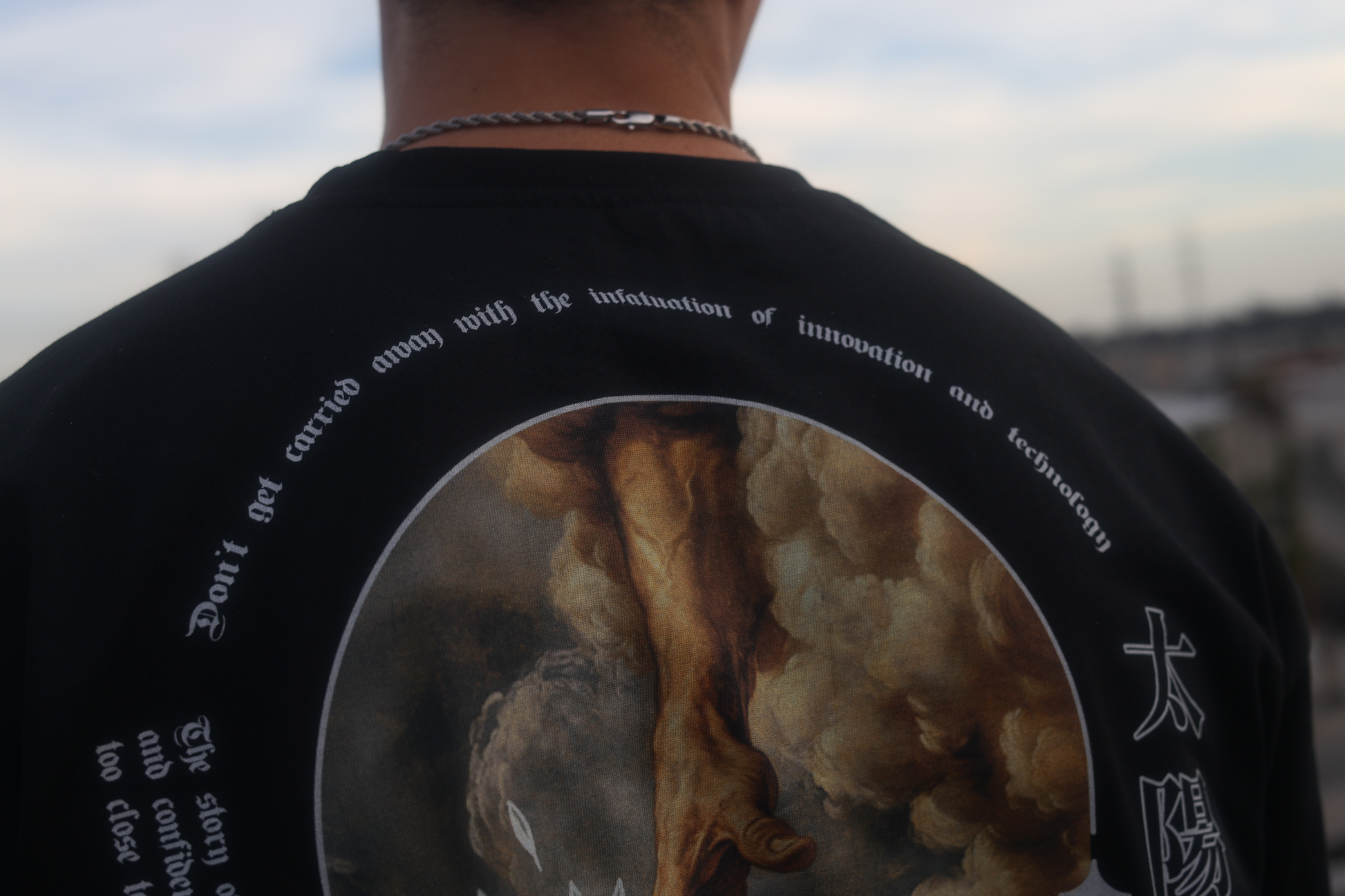 Closeup rear photo of a black oversize tee focused on the curved top text that says "don't get carried away with the infatuation of innovation and technology". Person is in the city.