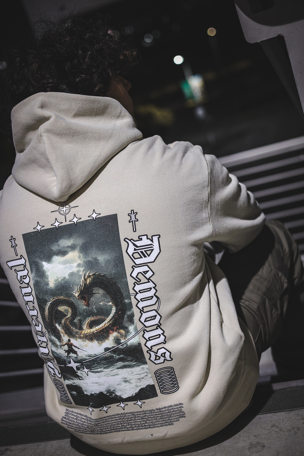 Rear photo of an ivory oversized drop shoulder hoodie consisting of a vertical rectangle graphic of a man with a sword in the water fighting a hydra in a storm. Large vertically angled gothic text of "Internal" on left and "Demons" on right. Set of four stars at the top/ bottom of vertical graphic, a single barbed wire on the top left/ right and interlocking circles on the bottom left/ right of the the vertical graphic. A story is written under the graphic. Prints are white with black outline.